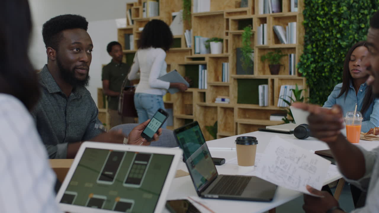 jóvenes empresarios afroamericanos se reúnen con un equipo de desarrolladores de software para diseñar aplicaciones móviles. colegas intercambian ideas creativas usando computadoras tablet. colaboran en la oficina de inicio.