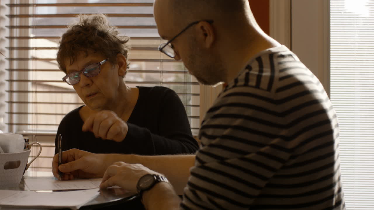 Senior mother sits at a kitchen table with adult son. They are reviewing, and signing papers ogether. Son hands pen over to mother, and she signs document. Both wear reading glasses. Warmly lit home.