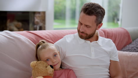 Cute-daughter-sitting-on-couch-with-father.-Cheerful-man-looking-at-small-girl