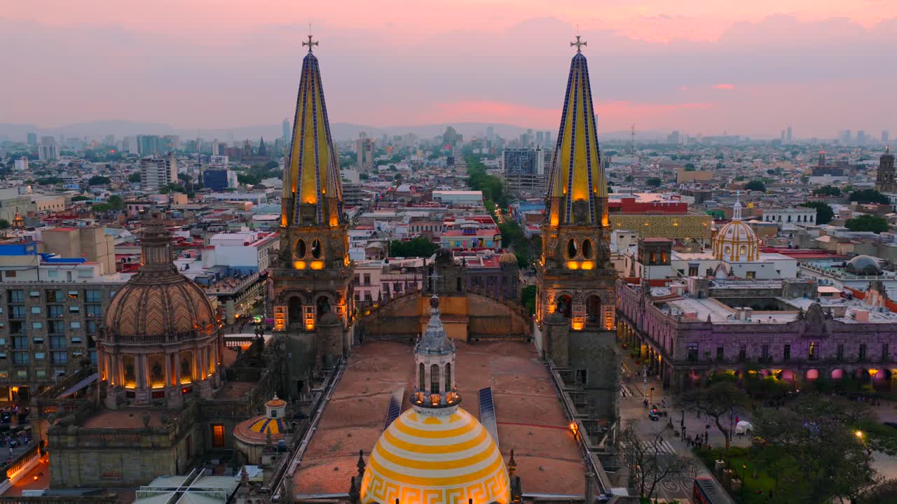Aerial dolly in flight toward illuminated dome and spires of Guadalajara Cathedral at sunset, flight reveals crowded Plaza de Armas below