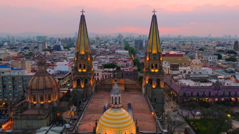 aerial dolly in flight toward illuminated dome and spires of guadalajara cathedral at sunset, flight reveals crowded plaza de armas below