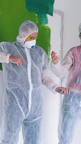 funny young couple wearing white protective suits and respirators dances near white wall with color strokes in light room