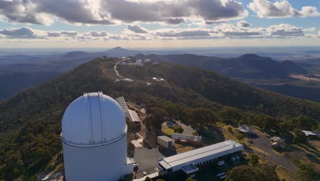 drone footage glides over siding spring observatory, revealing telescopes, research buildings, and forested hills under dramatic sunset lighting in rural australia
