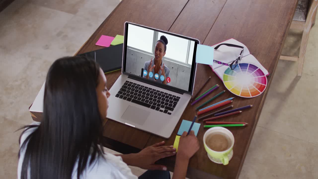 mujer caucásica usando una computadora portátil en videollamada con una colega y tomando notas