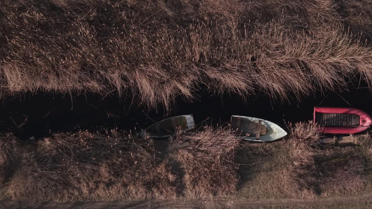 Veduta aerea dall'alto di piccole imbarcazioni in legno ormeggiate lungo uno stretto canale circondato da canne secche. L'acqua scura contrasta con la vegetazione dorata, creando una tranquilla scena di zona umida rurale