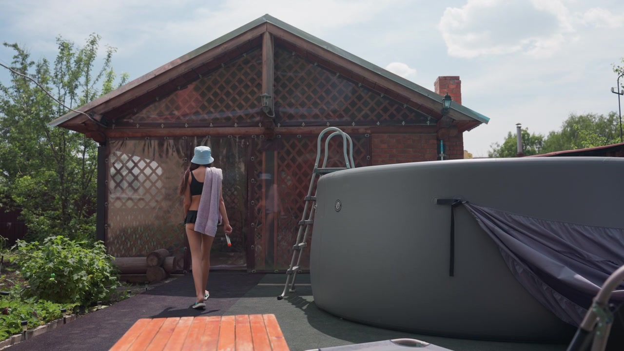 Una donna in costume da bagno nero e cappello da pescatore cammina verso una casetta di legno con traliccio, portando un asciugamano e tenendo un oggetto. L'ambiente esterno include una vasca idromassaggio, piante, cielo sereno e un'atmosfera calma.