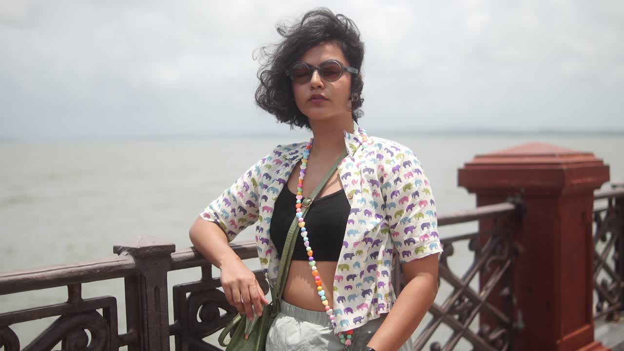 Medium shot moving closer to woman in sunglasses and open shirt leaning on seaside railing with wind in her hair