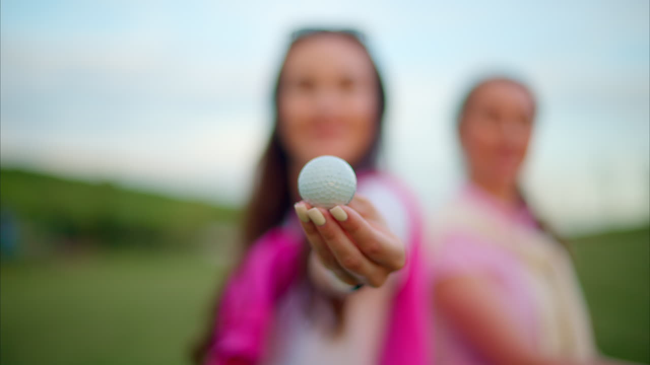 Close up of two women in white and pink clothes holding a white golf ball on the course