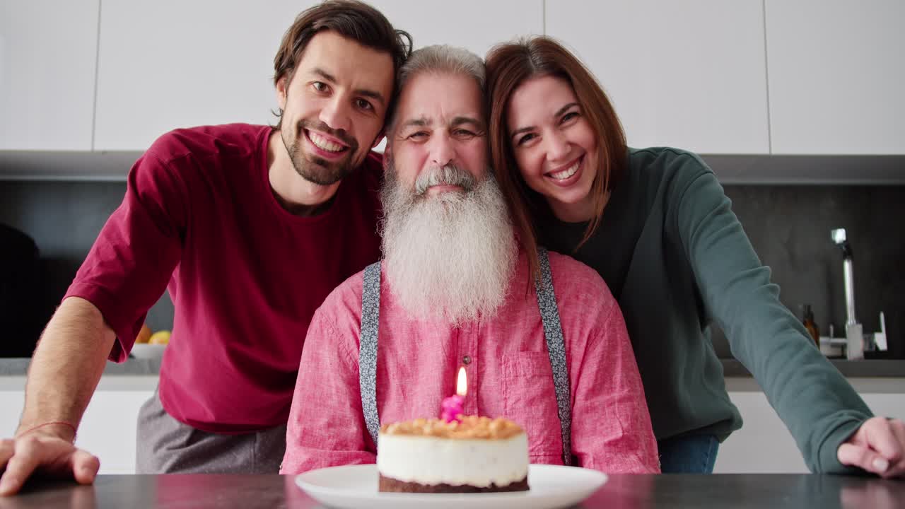 retrato de un hombre mayor feliz con cabello gris con una barba exuberante en una camisa rosa que se sienta en la mesa y delante de él es un pastel con una vela encendida al lado de sus hijos adultos un hombre moreno con paja y una chica morena en la cocina en un apartamento moderno