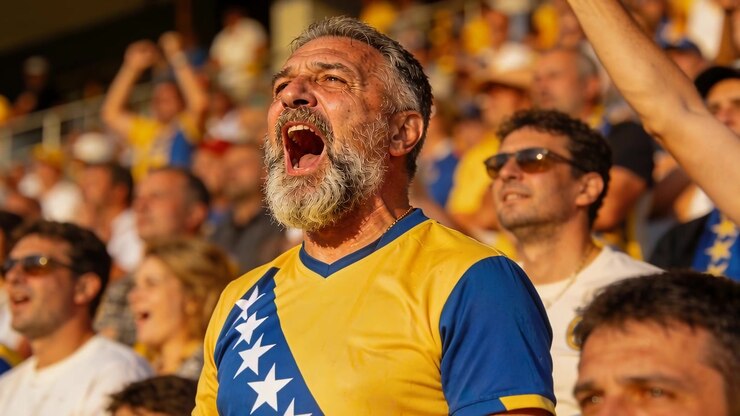 Tifoso anziano barbuto che urla reagendo a un'azione di gioco dal vivo sugli spalti, indossando una maglia giallo-blu con stelle