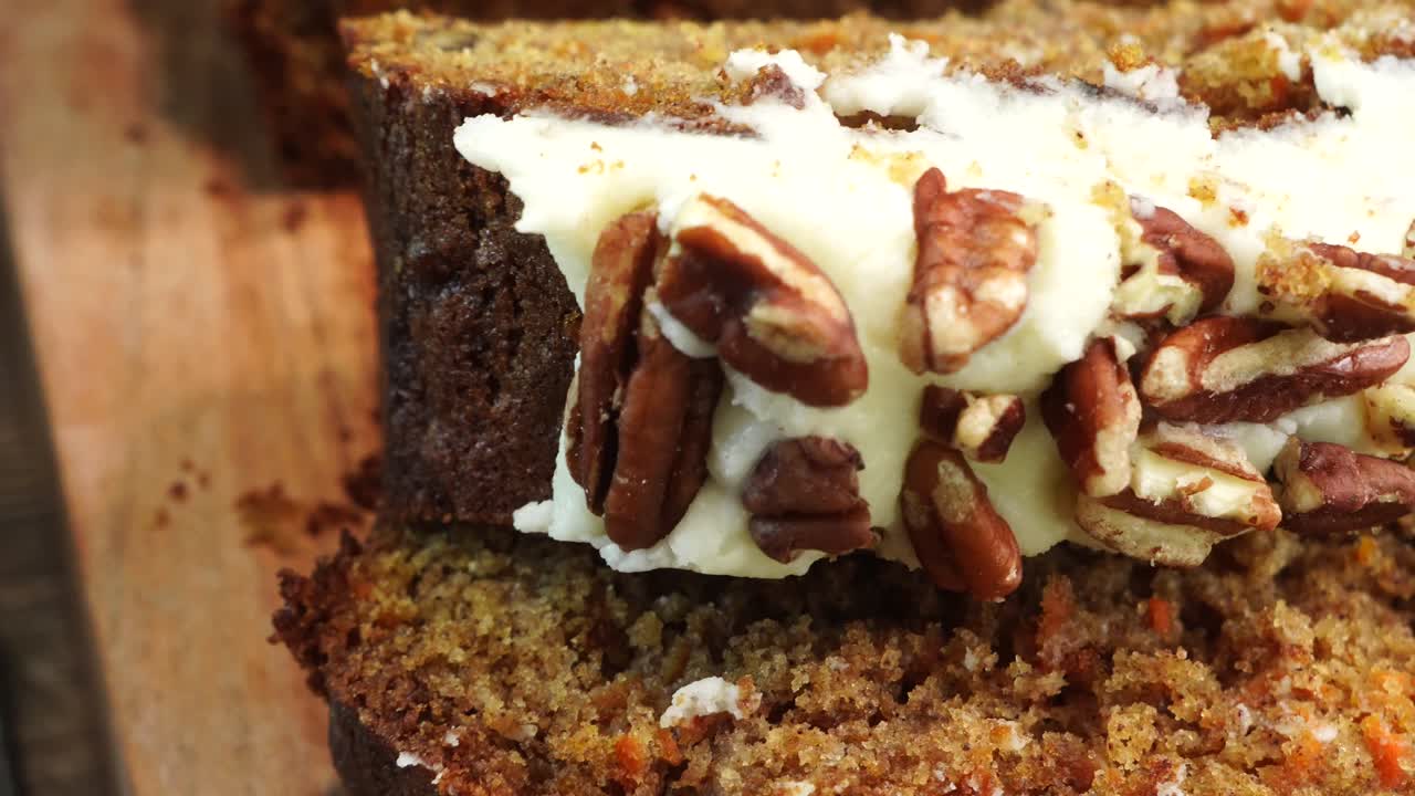 Baking a carrot cake loaf with cream cheese icing and topped with roasted salted pecans. Making a Autumn dessert with carrots, flour, sugar, nuts and frosting. Panning across the carrot cake loaf.