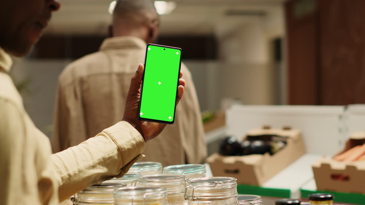 comerciante afroamericano sosteniendo un teléfono con pantalla verde en la tienda de comestibles