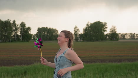 side view of excited woman in denim dress running joyfully along straight asphalt road holding colourful pinwheel against cloudy sky hair tousled by summer breeze
