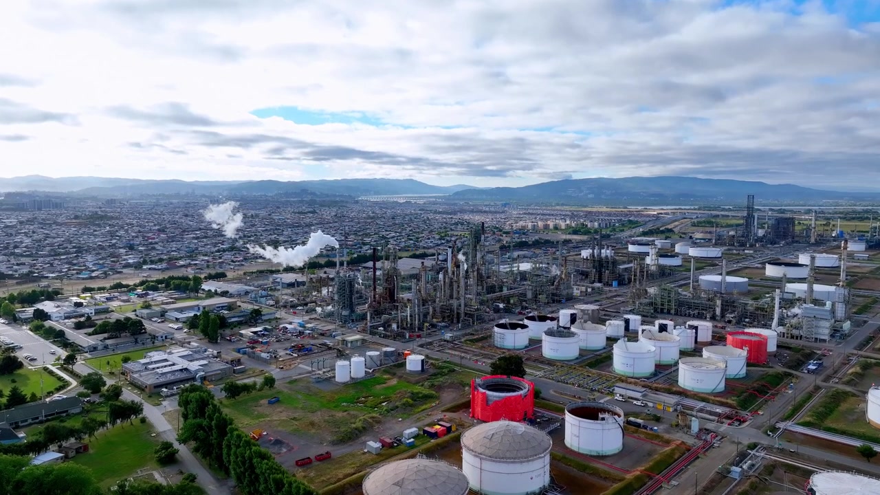Vuelo frontal sobre un vasto complejo de refinería de petróleo con torres de destilación, vapor y tanques de almacenamiento, junto a una densa ciudad residencial y una cordillera que se extiende hasta el horizonte. Hualpén, Biobío, Chile.