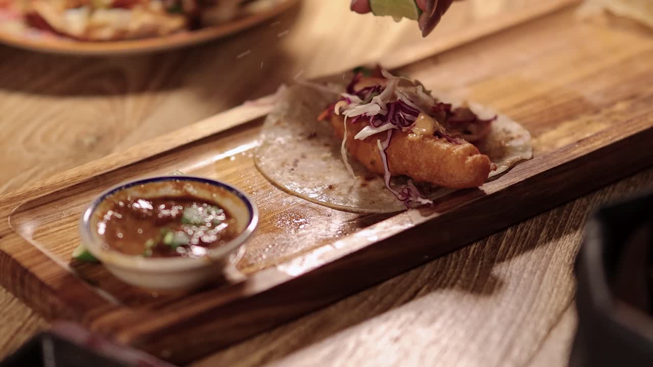 A hand squeezes fresh lime over a crispy fish taco with slaw on a soft tortilla, served on a wooden board under warm, ambient lighting