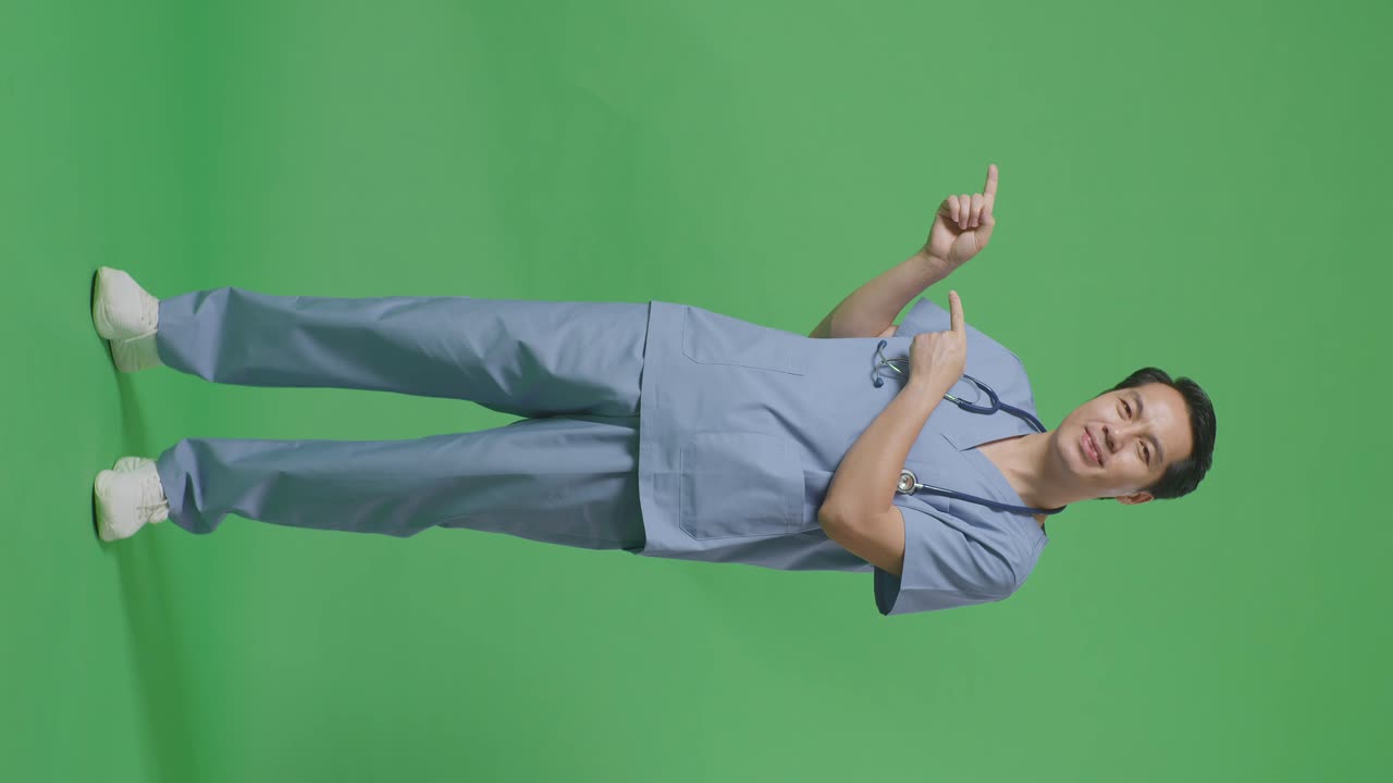 스테토스코프를 들고 있는 아시아 남성 의사의 몸 전체가 미소 짓고 병원의 녹색 화면 배경에서 으로 가리키고 있습니다.