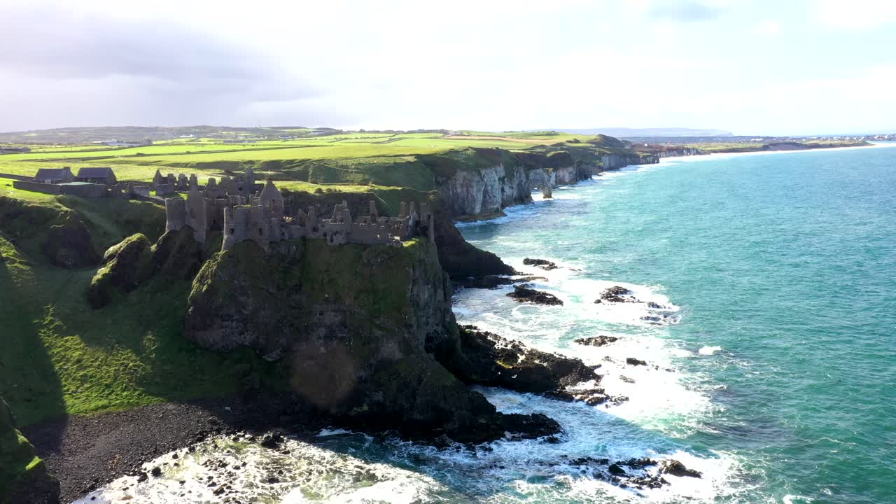 Slow drone establishing dolly along Irish coastline as waves crash against lush green cliffs below Dunluce castle