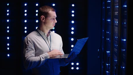 Young-man-holds-device-in-hand-and-looks-at-screen-inspects-equipment-or-hardware-rack.-Male-programmer-working-with-laptop-and-supporting-service-while-standing-in-data-center-spbas.