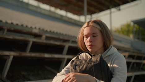 close-up of lady in deep thought seated alone on empty stadium bleachers, hugging her bag with a reflective expression and distant gaze, sunlight softly illuminates her as she contemplates quietly