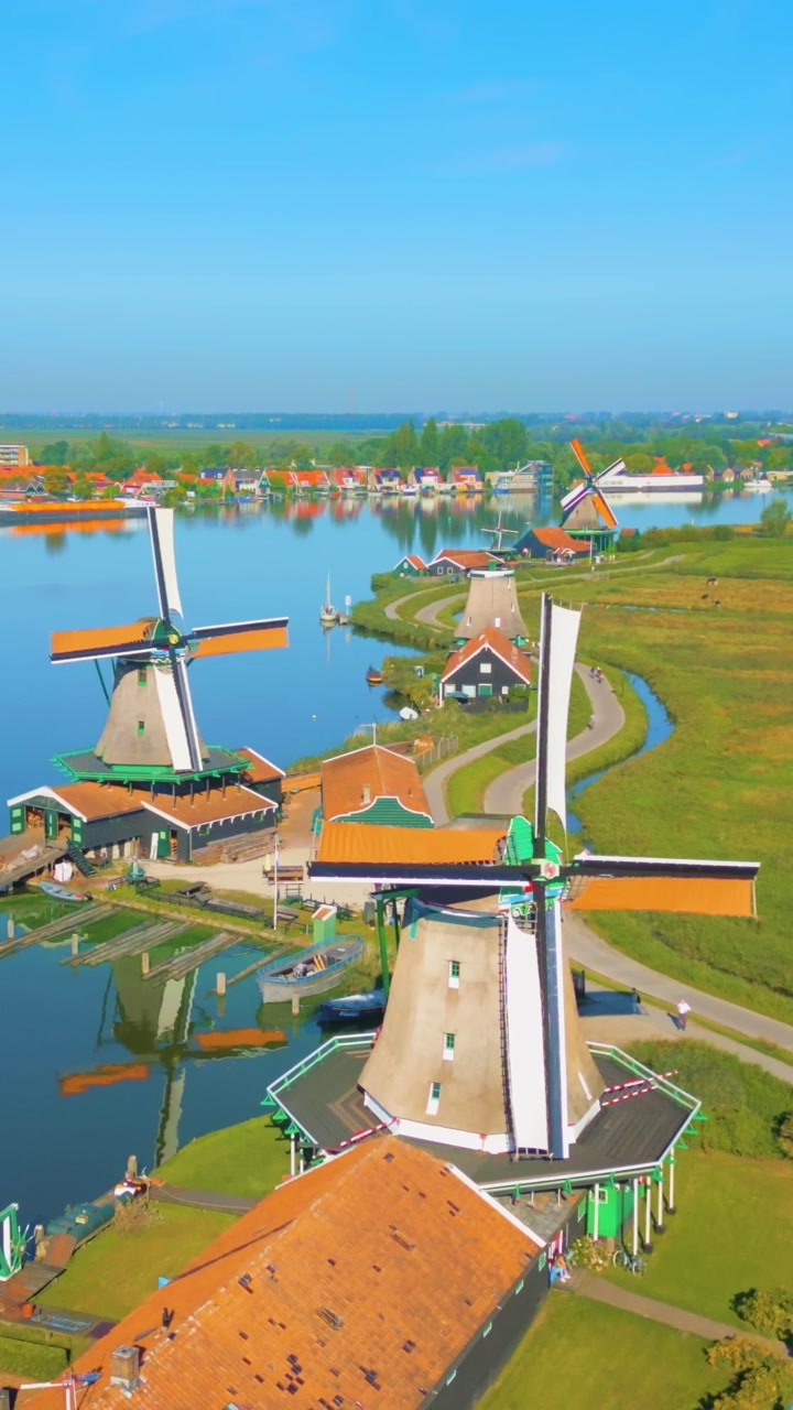 Wide aerial view of numerous historic windmills and traditional Dutch houses at Zaanse Schans. The picturesque village landscape features winding paths, green fields, and tranquil canals under a br...