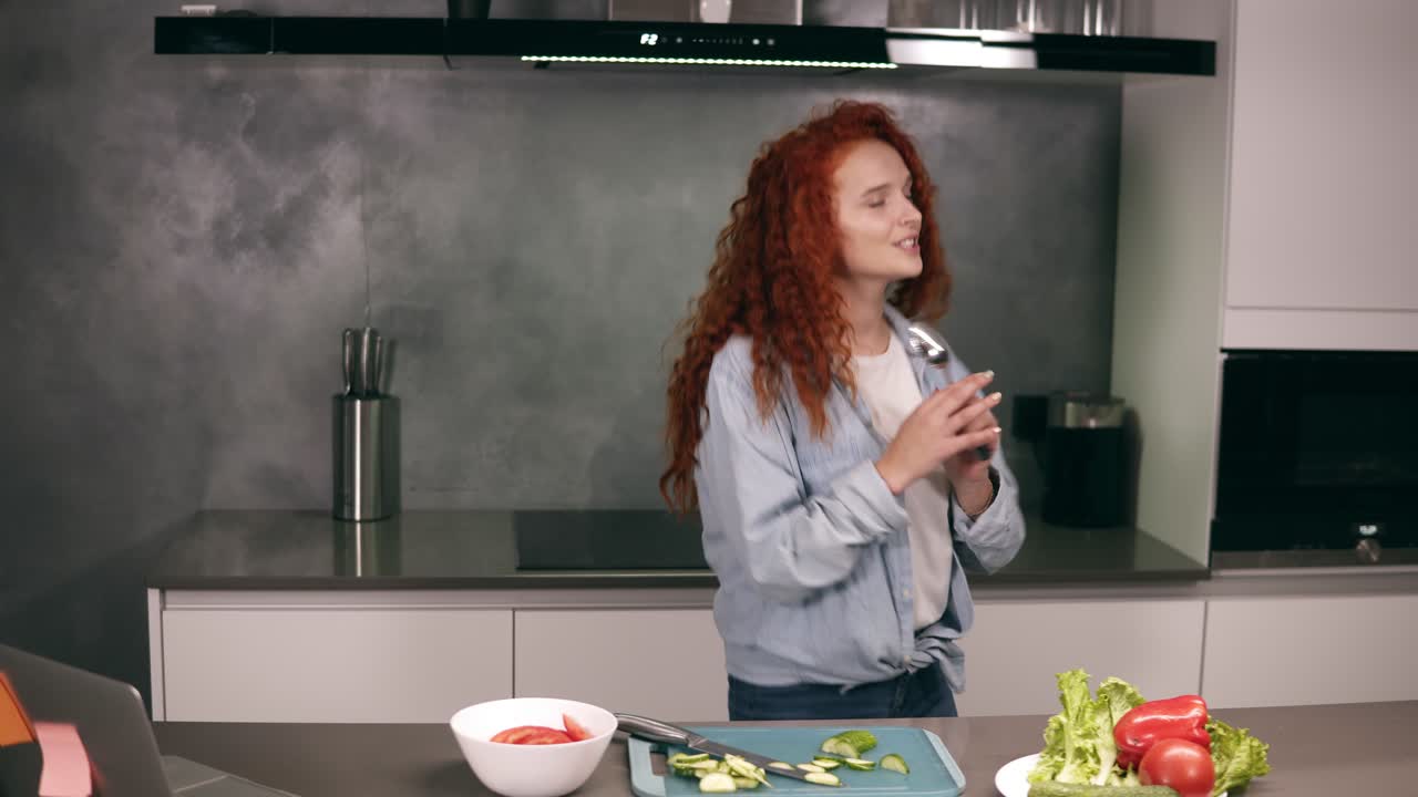 Happy active redhead young woman holding spoon as a microphone singing songs and dancing while cooking in kitchen, carefree