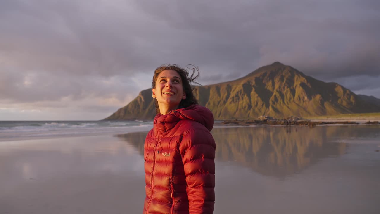 cámara lenta, una hermosa mujer joven admirando la vista y sonriendo en la playa de flakstad frente a una hermosa puesta de sol dorada, islas lofoten, noruega
