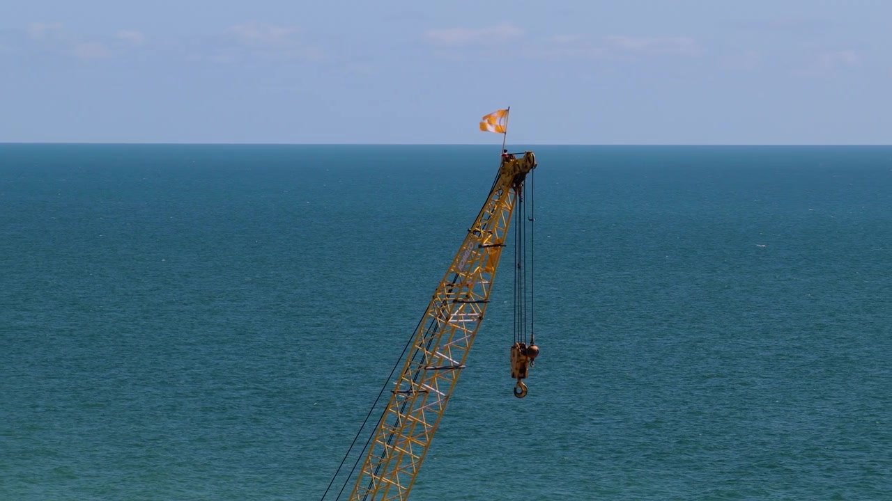 Yellow lattice boom crane with hanging hook and flag rises against a calm blue ocean and clear sky.