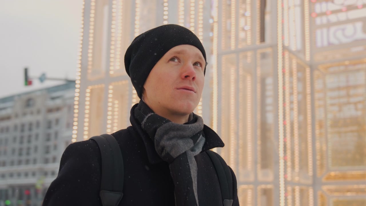 Close up of man wearing winter clothing standing outdoors in urban environment. subject is dressed in black coat gray scarf and beanie with snowflakes visible on his attire as man looks