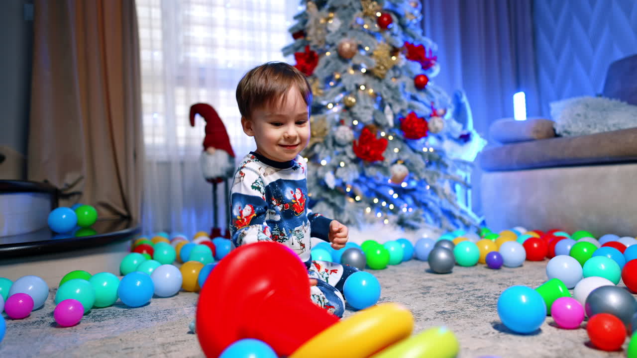 Cheerful baby sitting near the beautiful Christmas tree. Smiling toddler builds a pyramid and throws it on the floor.