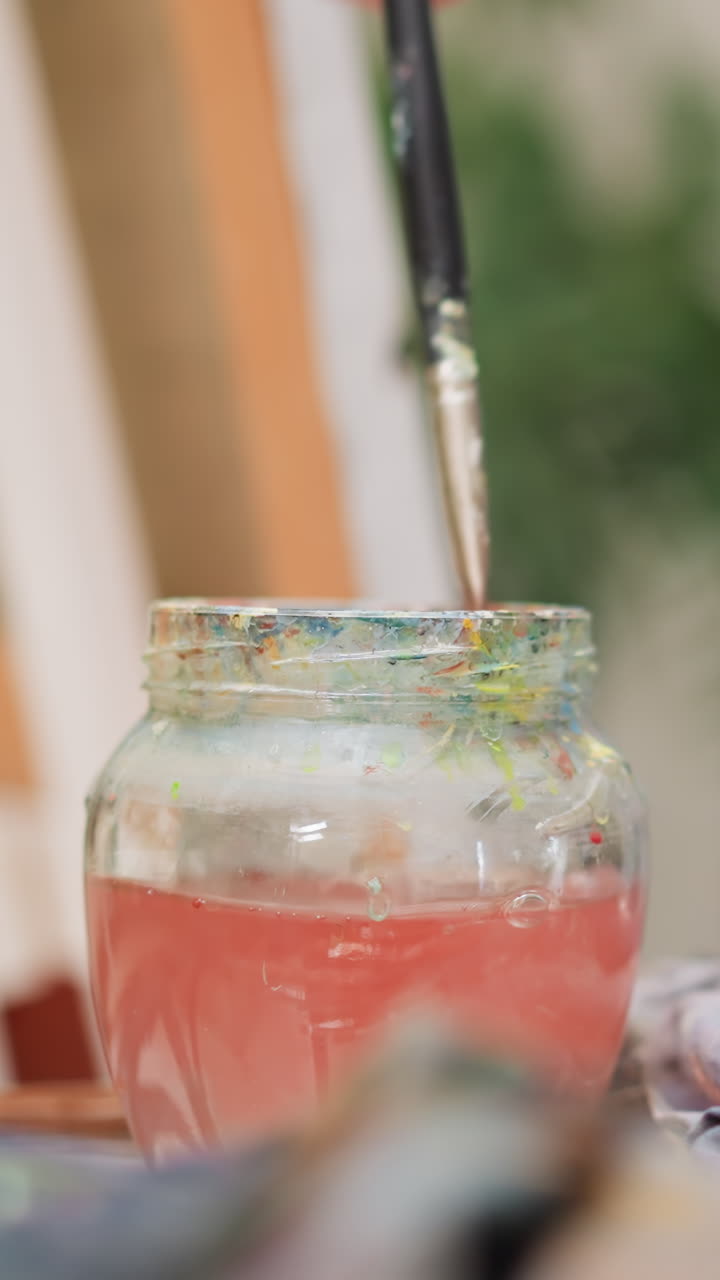 el artista lava el pincel en agua vertida en un frasco de vidrio de cerca. el maestro de arte limpia el pincil del pigmento rojo en la mesa de la tienda. el hombre creativo usa una herramienta de dibujo