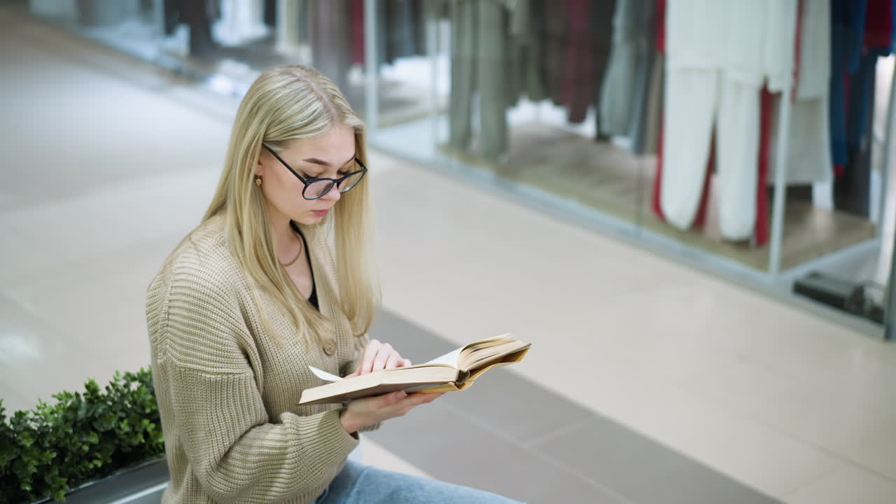 el estudiante con gafas de google se centra en el libro, volteando las páginas pensativamente con una flor decorativa verde detrás, la ropa visible a través del vidrio de la tienda