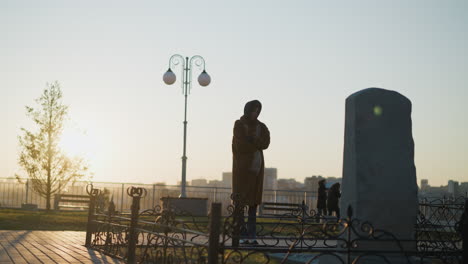 a tearful woman wearing a coat and a scarf stands in front of a stone monument in a park during sunset. moment of sorrow and contemplation, with the warm glow of the setting sun casting long shadows