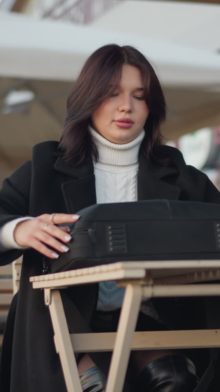 Lady seated outdoors at mall unzips bag on wooden table in modern urban setting, stylish outfit includes black coat and white sweater, background features tables, chairs, and blurred storefronts