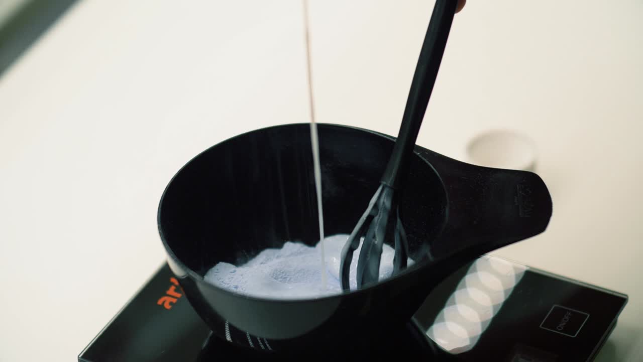 Pouring hair dye ingredients into black plastic bowl and mixing custom color for salon customer. Exact measurements essential for proper color results. Customized client hair treatment in spa.