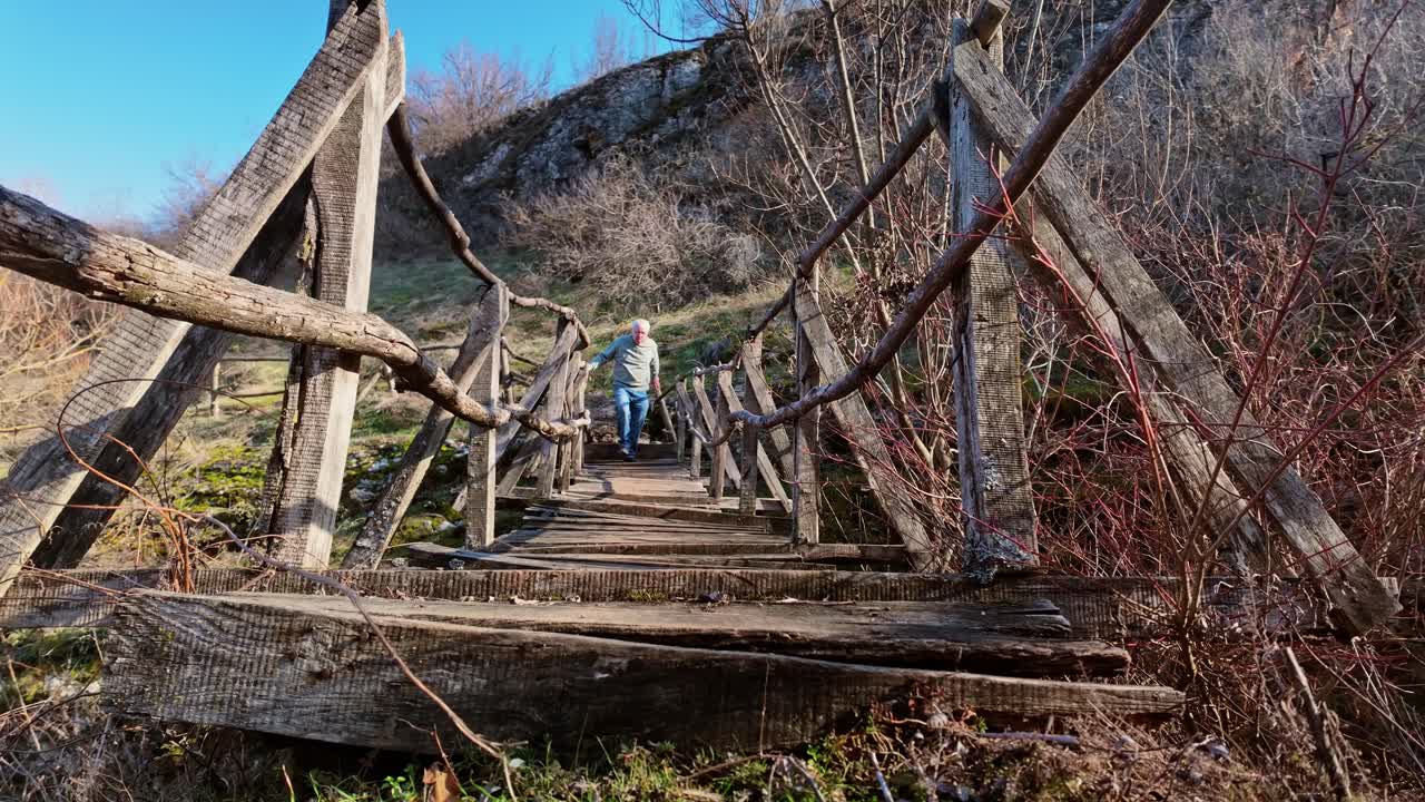 Man grips log rail supports as strides over a risky wooden hike trail footbridge.