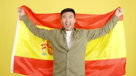 excited asian man cheering with spanish flag on yellow background