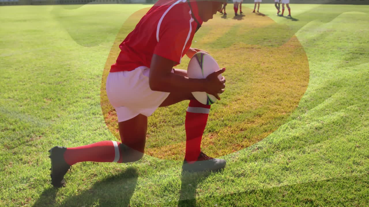 Kneeling on field, rugby player holding ball with teammates in background