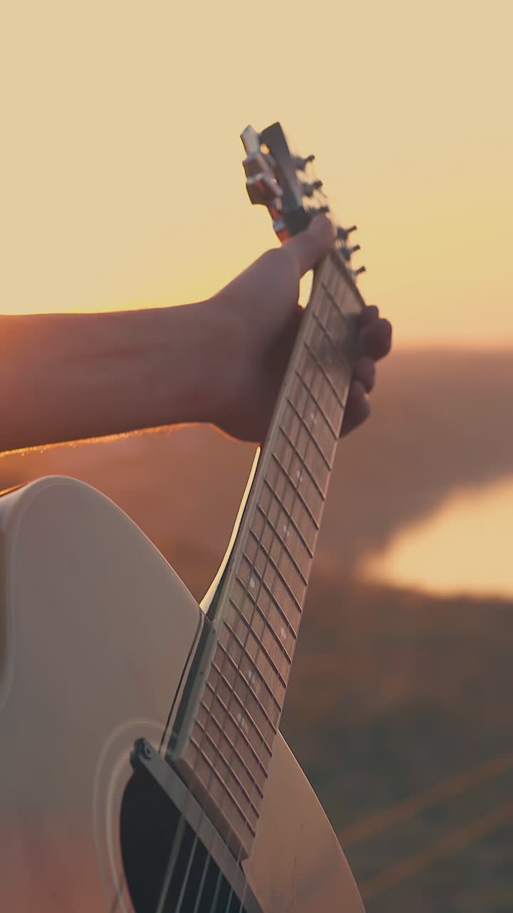 young man plays music with guitar in tourist camp against calm river and back sunset in warm autumn evening closeup