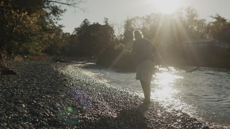 A-homeless-woman-in-rubber-boots-walks-along-the-edge-of-a-lake,-looking-for-something-on-the-shore.