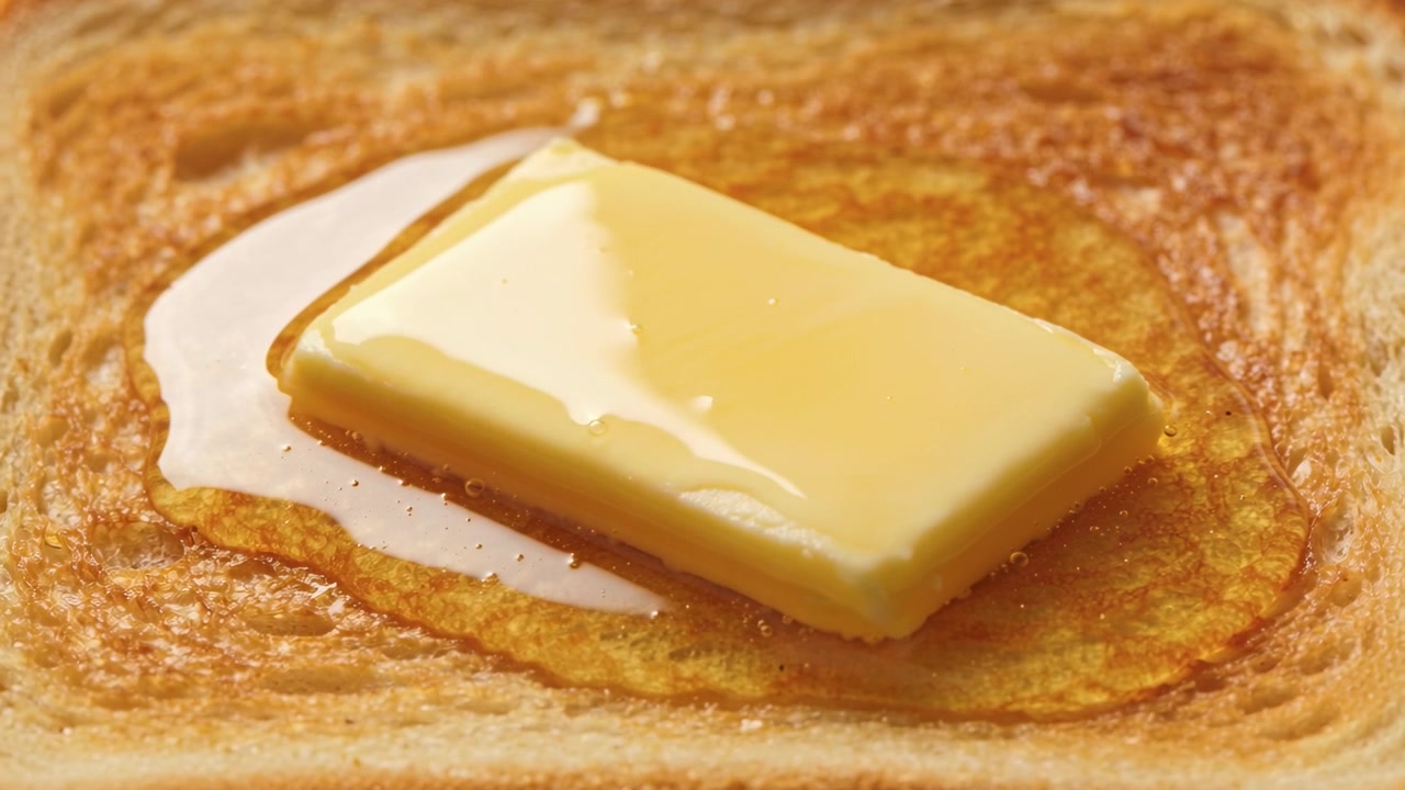 Morceau de beurre fondant s'étalant et noircissant sur un toast en gros plan sous l'effet de la chaleur, révélant la texture de la mie.