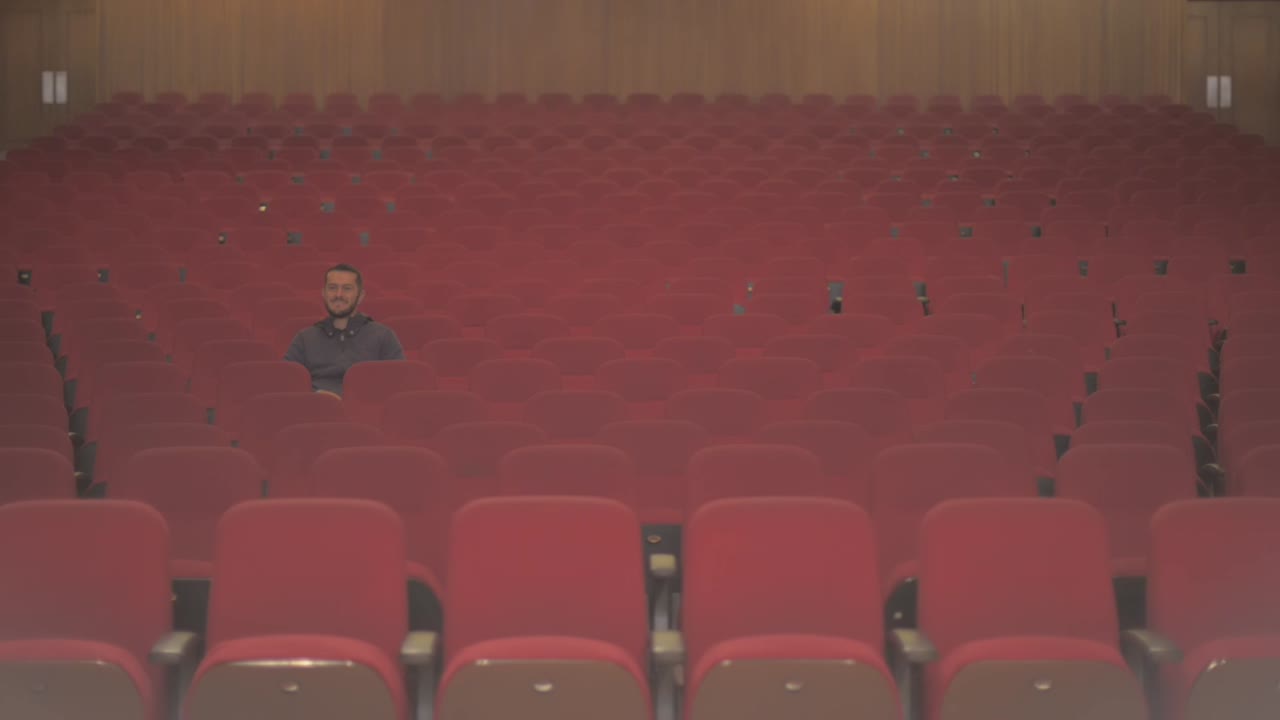A tall white guy is in an empty theatre. He is the only one watching the comedy show and he starts to laugh while he enjoys what he sees on stage