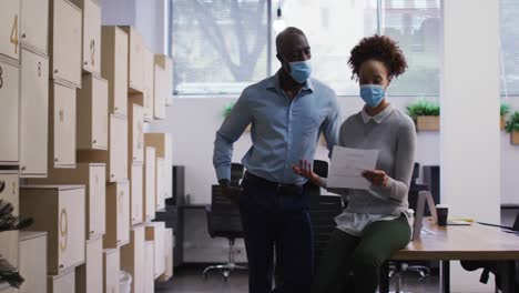 diverse male and female business colleagues in face masks talking, woman holding document in office