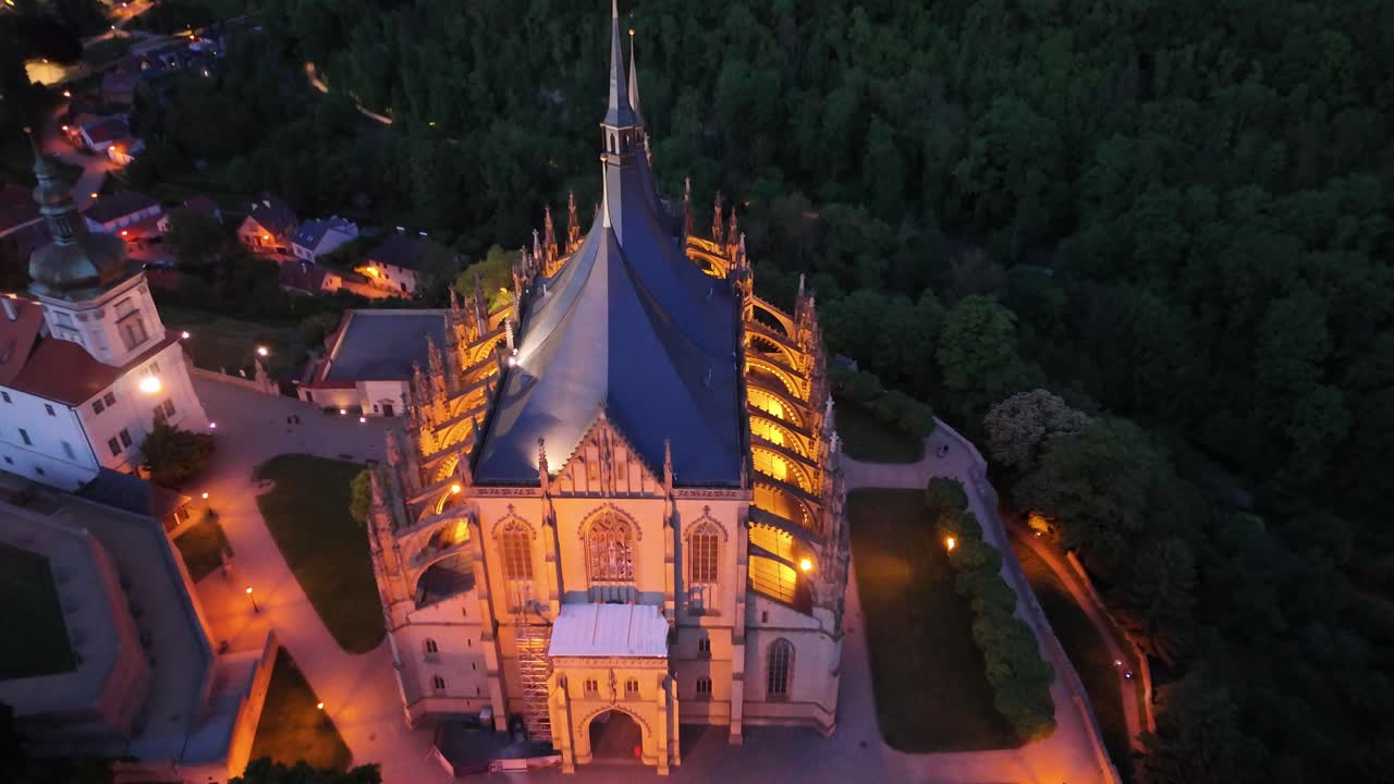 Aerial spin around illuminated Saint Barbara Cathedral at twilight, showing urban surroundings