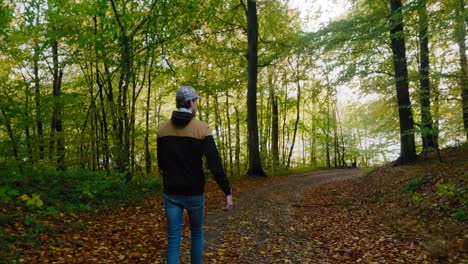 Joven-Con-Gorra-Y-Chaqueta-Camina-Por-Un-Pequeño-Sendero-En-El-Bosque-De-Gyllebo-En-La-Tarde-De-Auyumn,-Österlen-Suecia---Seguimiento-De-Tiro-Ancho-Medio-Desde-Atrás