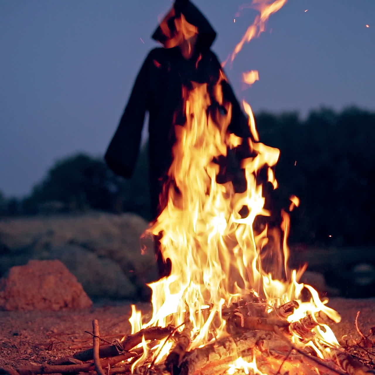 Horrible black silhouette behind the bright flame outdoors. Vivid flame from fire in the evening and witch in black costume waving arms making fear.