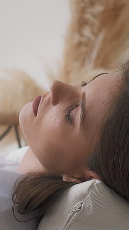 sad tired young woman with dark hair and bare knees in white shirt sits in armchair with soft pillows in light room at home close side view slow motion