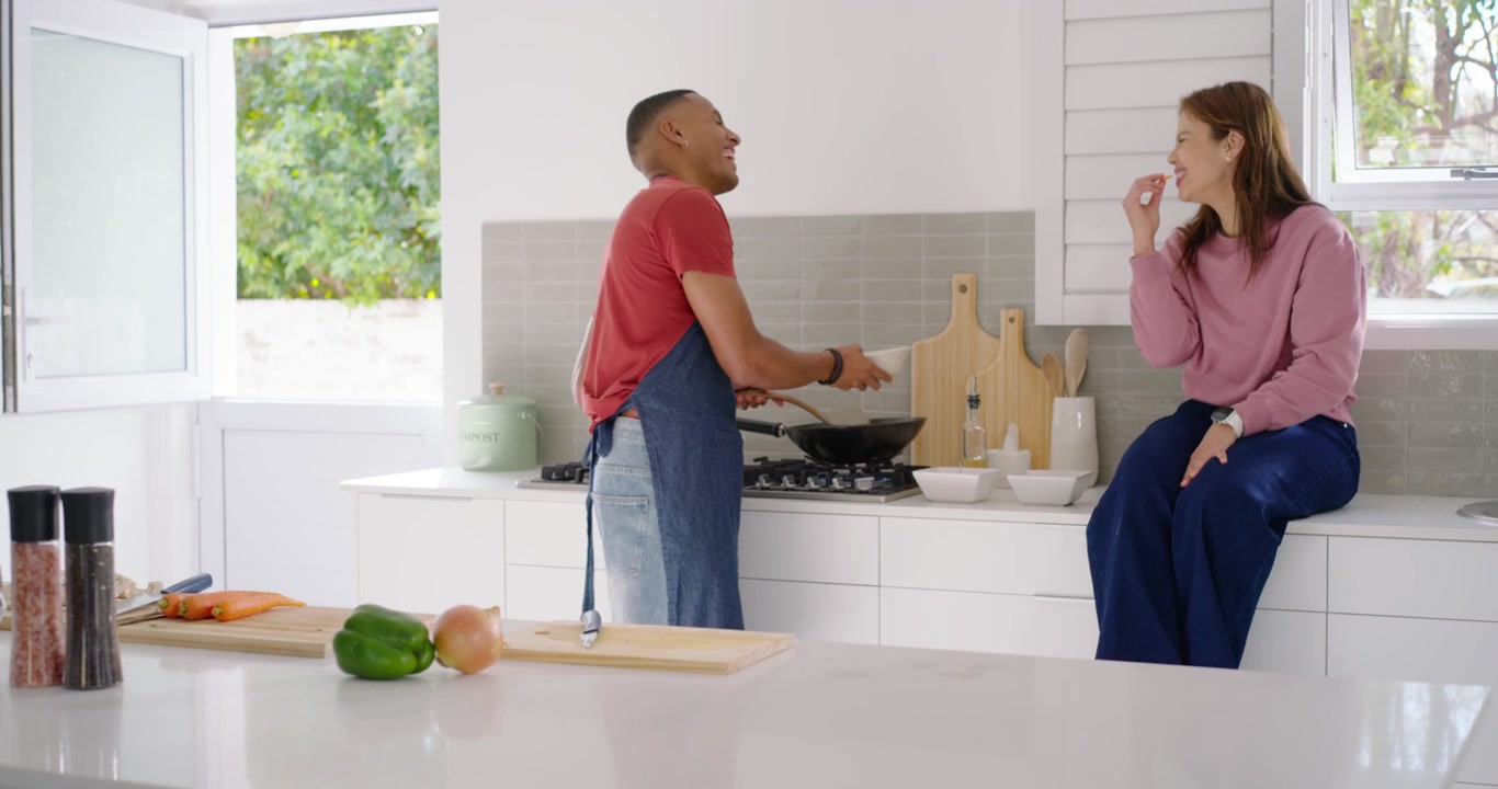 Coppia multietnica che prepara un pasto da condividere, l'uomo mescola la padella con una spatola mentre la donna porge l'olio.