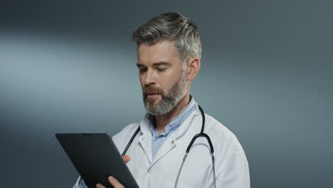 apuesto médico de bata blanca usando un dispositivo de tableta, tocando y desplazándose en el fondo de la pared gris