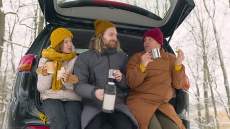 tres amigos bebiendo y comiendo sándwich mientras estaban sentados en el maletero del camión durante un viaje por carretera de invierno