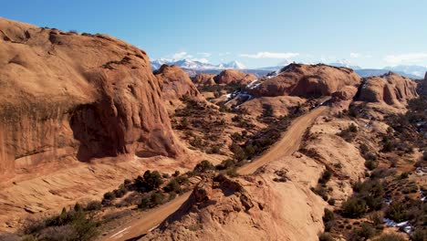 Eine-Hochfliegende-Drohne-Schoss-über-Eine-Abgelegene-Unbefestigte-Straße,-Die-Durch-Das-Weite-Und-Einzigartige-Wüstenland-In-Der-Nähe-Von-Moab,-Utah,-Führt,-Mit-Den-Schneebedeckten-Rocky-Mountains-In-Der-Ferne